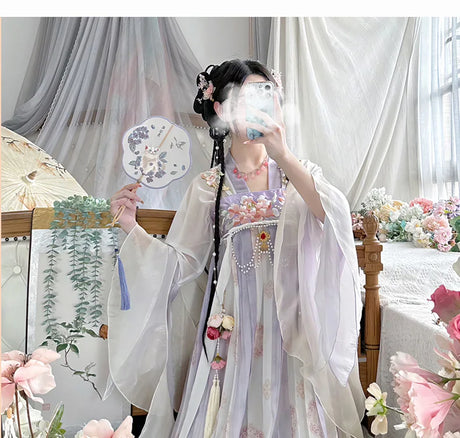Woman in a vintage Chinese Hanfu dress holding a decorative fan, showcasing traditional elegance with floral decor in background.