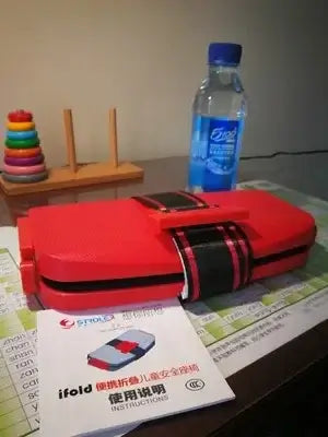 Ifold Portable Baby Car Seat in red with instruction manual, a water bottle, and toys on a table.