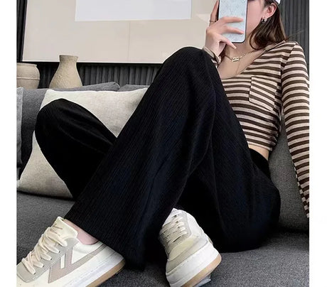 Woman wearing Ice Silk Pleated Maternity Pants in black, paired with a striped top and sneakers, seated on a couch.