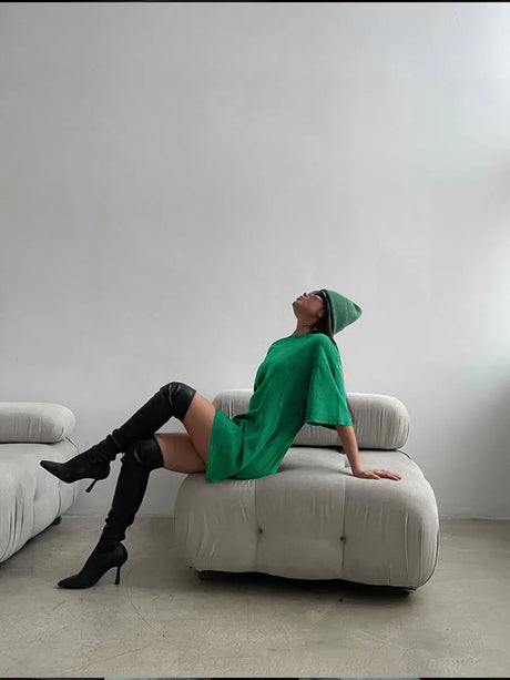 Model in a green oversized t-shirt dress and knee-high boots, lounging on a modern sofa in a minimalist setting.