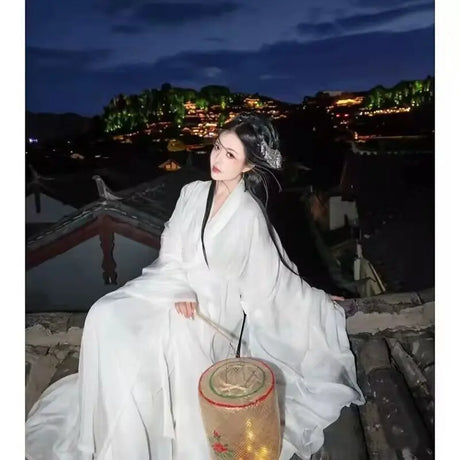 Elegant woman in traditional white Hanfu dress sitting on rooftop at night with lantern, showcasing Chinese cultural beauty.