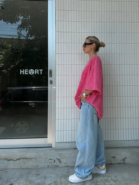 Woman wearing a pink oversized shirt and baggy jeans, standing outside a store with 'HEART' signage.