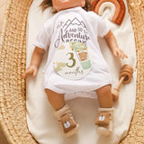 Baby doll wearing a white onesie with '3 months' milestone and bear pattern, placed in a cozy basket.