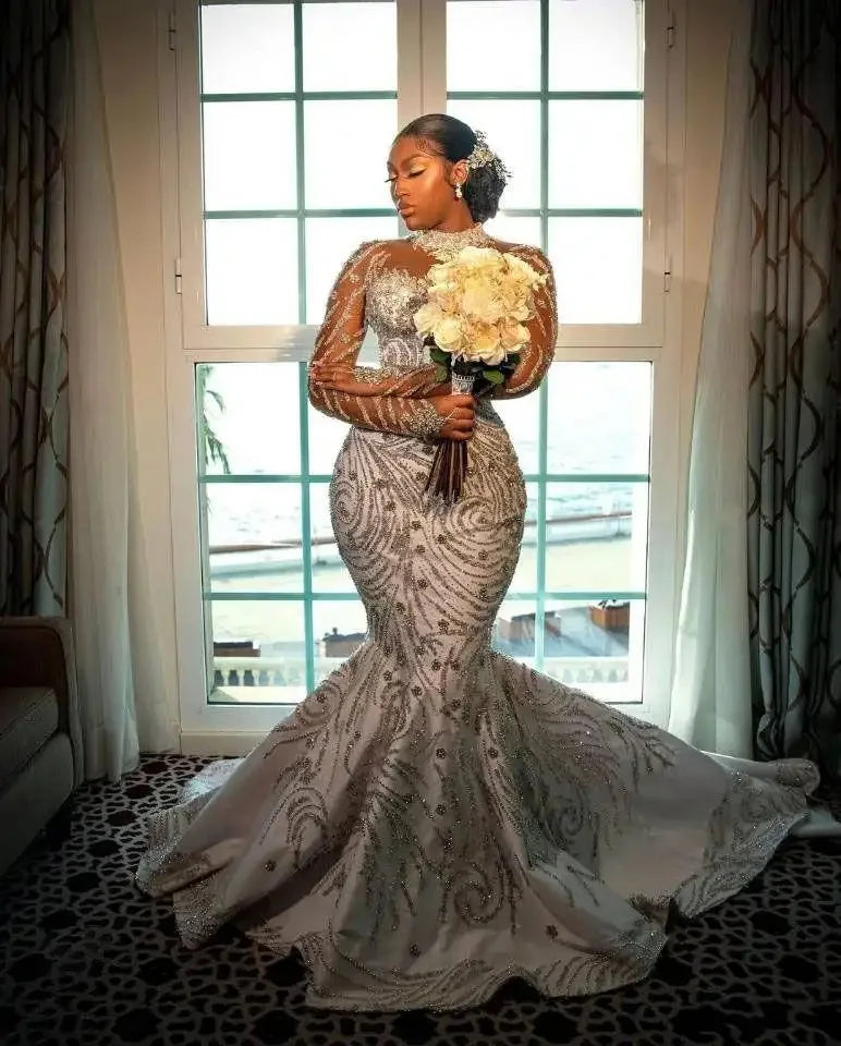 Luxury beaded mermaid wedding dress with sheer sleeves, model holding bouquet, elegant backdrop with window.