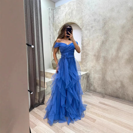 Woman in a stunning blue off-the-shoulder evening gown with layered ruffles standing in a chic indoor setting.