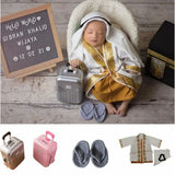 Baby boy dressed in traditional clothing beside a mini suitcase and accessories, showcasing creativity and comfort.