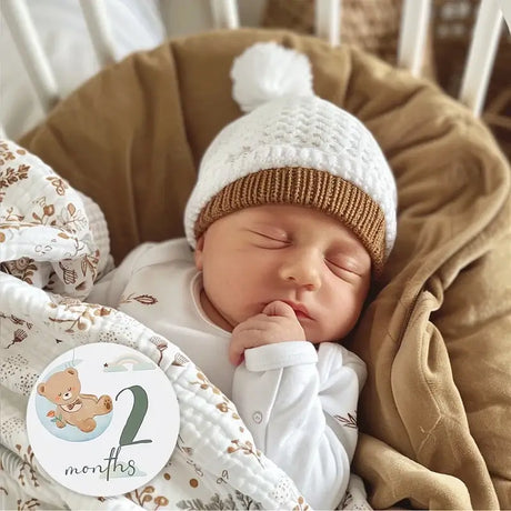 Sleeping baby in a cozy hat with a milestone card showing '2 months', surrounded by soft blankets and a peaceful atmosphere.