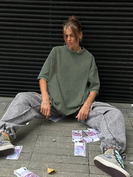Model wearing a loose, oversized casual t-shirt paired with baggy pants and sneakers, sitting on the ground surrounded by dollar bills.