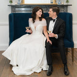Bride in sheer VG73 wedding jacket with groom, both enjoying drinks on a blue velvet sofa.