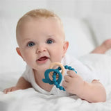 Smiling baby lying on a bed, holding a blue silicone teether and wooden ring, showcasing safe and non-toxic materials.