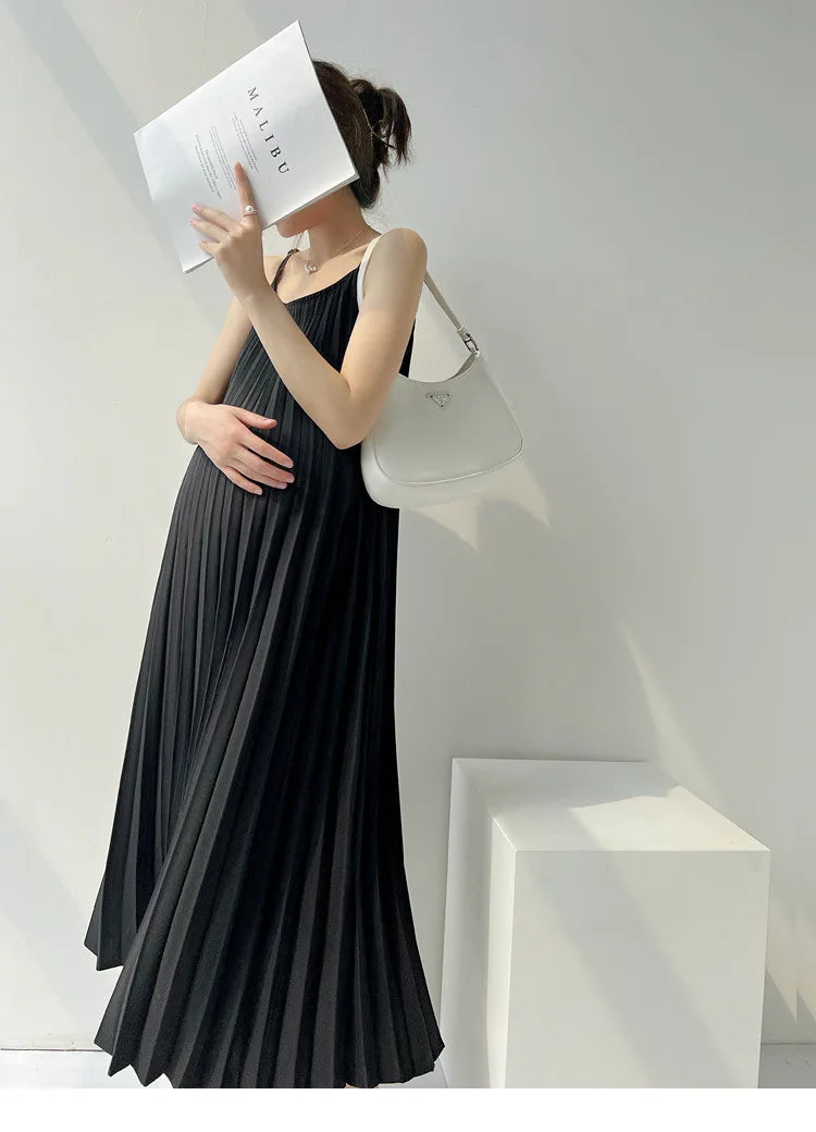 Pregnant woman wearing a pleated black chiffon maternity dress, holding a book, and accessorized with a white handbag.