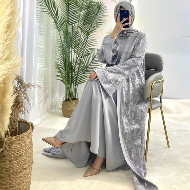 Elegant woman in a grey Islamic kaftan and hijab, sitting in a modern setting with plants and stylish decor.