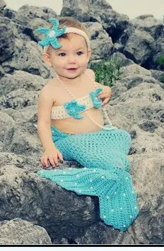 Baby wearing a blue mermaid costume with a flower headband, sitting on rocks in a natural setting.