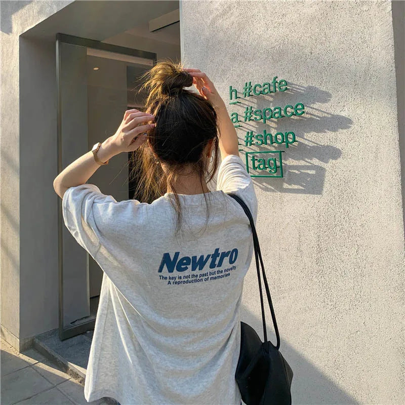 Model wearing oversized gray tee with 'Newtro' print, standing by a cafe sign.