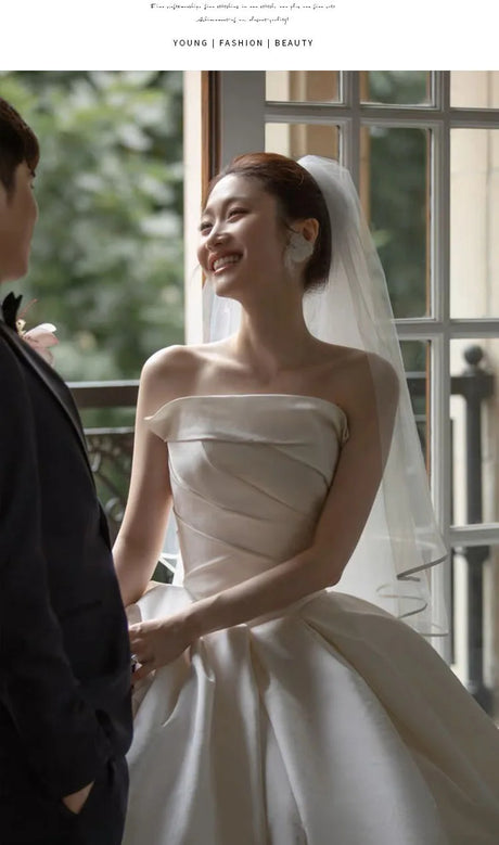 Bride wearing an off-shoulder satin wedding dress with a veil, smiling at her partner indoors.