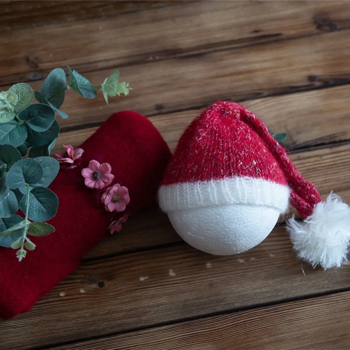 Newborn stretch knit mohair in a festive red and white hat on a wooden background, surrounded by flowers and greenery.