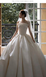 Elegant bride in a satin off-shoulder wedding gown standing by a window, showcasing a voluminous skirt and delicate veil.