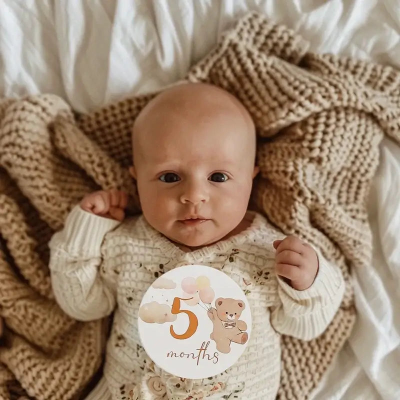Baby girl laying on a cozy blanket holding a 5-month milestone card featuring a bear pattern.