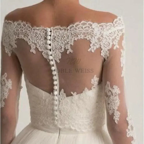 Lace off shoulder bridal jacket by Noble Weiss, featuring long lace sleeves and elegant button detailing for a stunning bridal look.