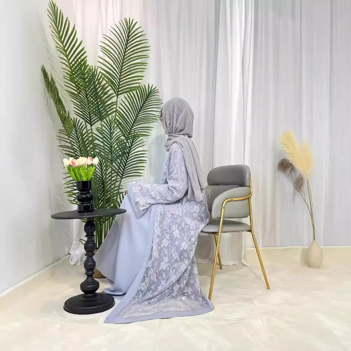 Elegant Muslim woman in gray Dubai Moroccan Kaftan, seated by a table with flowers and greenery, showcasing Islamic fashion.