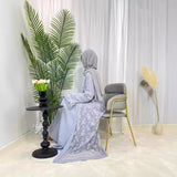 Elegant Muslim woman in gray Dubai Moroccan Kaftan, seated by a table with flowers and greenery, showcasing Islamic fashion.