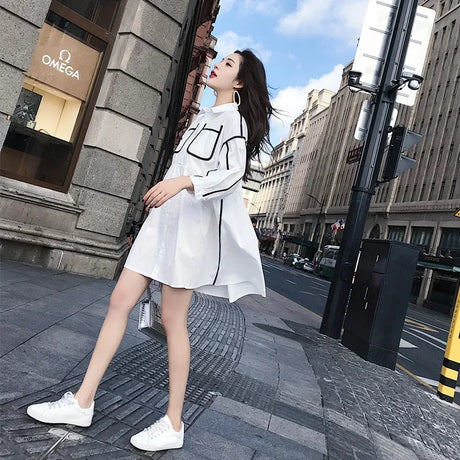 Model wearing a solid color maternity shirt dress in white, styled with sneakers, walking on an urban street.