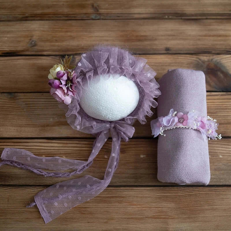Newborn photography props featuring a purple bonnet, wrap, and floral headband on a wooden surface.