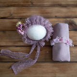 Newborn photography props featuring a purple bonnet, wrap, and floral headband on a wooden surface.