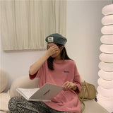 Woman in an oversized pink Harajuku tee and beret, sitting with a book in a stylish indoor setting.