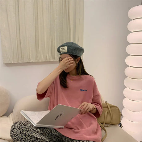 Woman in an oversized pink Harajuku tee and beret, sitting with a book in a stylish indoor setting.