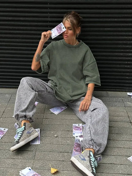 Women lounging in a casual outfit, featuring a loose olive green t-shirt and gray pants, styled with patterned sneakers.