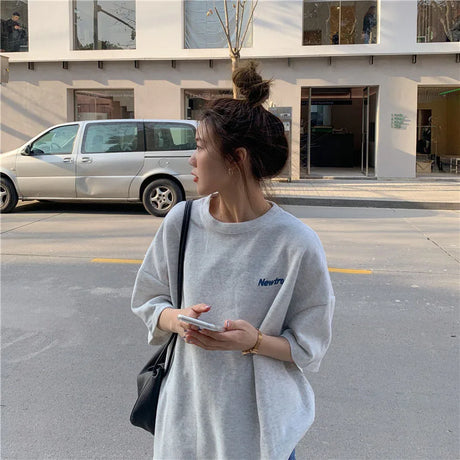 Woman in oversized gray Harajuku streetwear tee, holding a phone, standing outside.