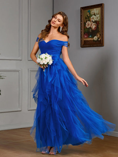 Model wearing a vibrant blue off-the-shoulder gown with layered tulle, holding a bouquet in an elegant setting.