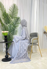 Elegant woman in grey Moroccan kaftan and hijab, seated by a table with decorative plant and backdrop.