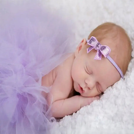 Newborn baby girl in purple infant tutu skirt and headband sleeping on cozy surface, perfect for photography accessories.