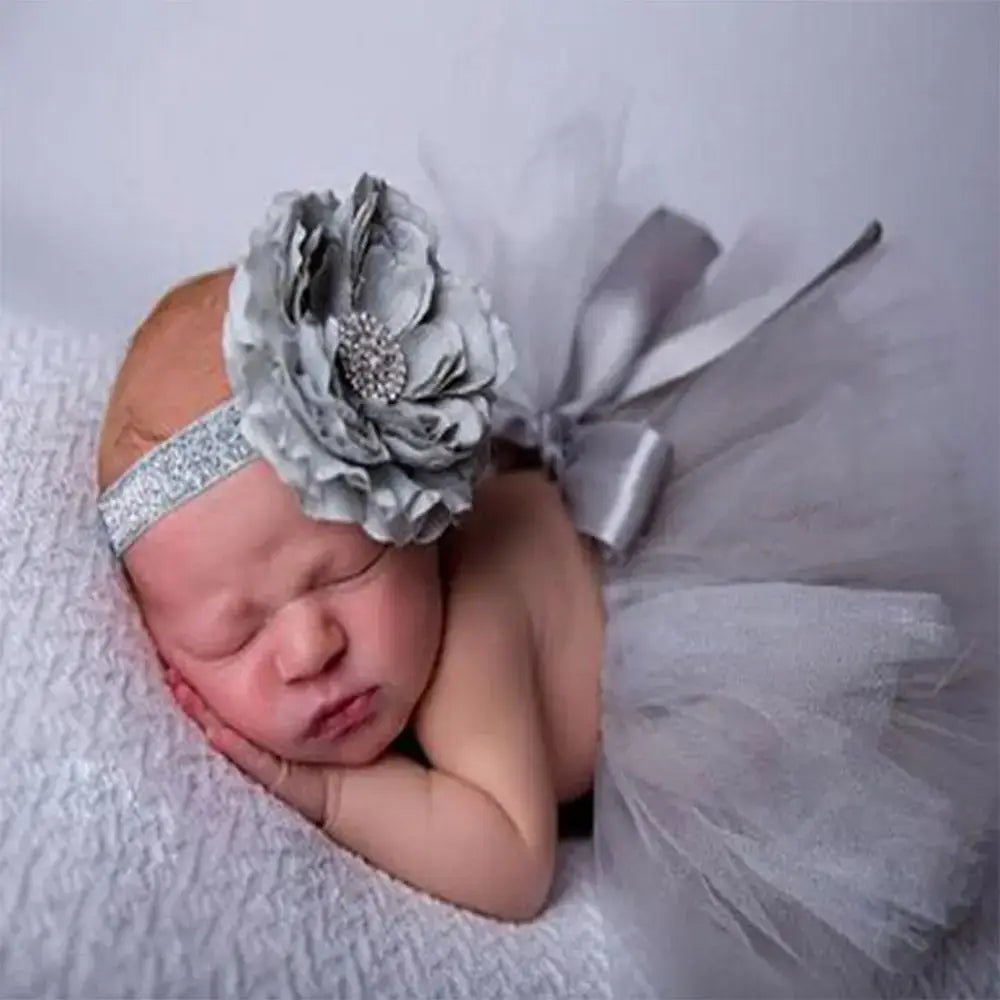 Newborn baby girl in a gray tutu skirt and matching headband, sleeping peacefully on a soft white blanket.